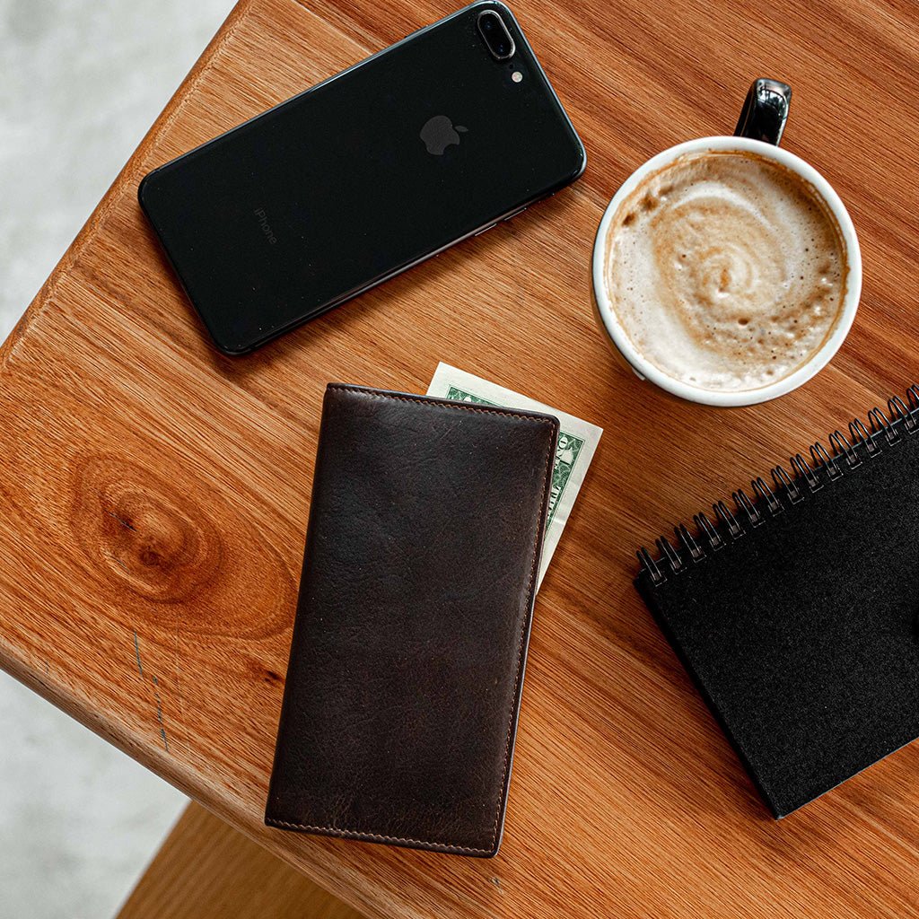 Dark brown leather long wallet on a wooden table with a phone, coffee and notebook