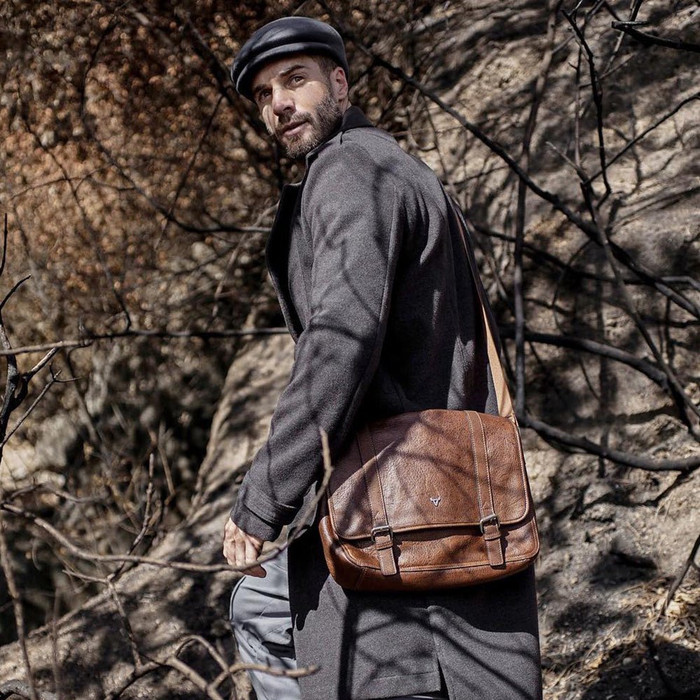 Man in a flat cap carrying a brown leather messenger bag outdoors
