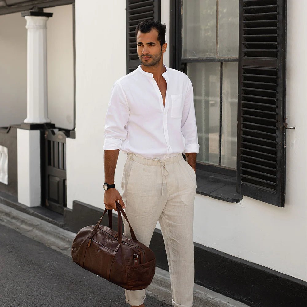 Man in white linen walking with a brown leather duffel bag on a city street