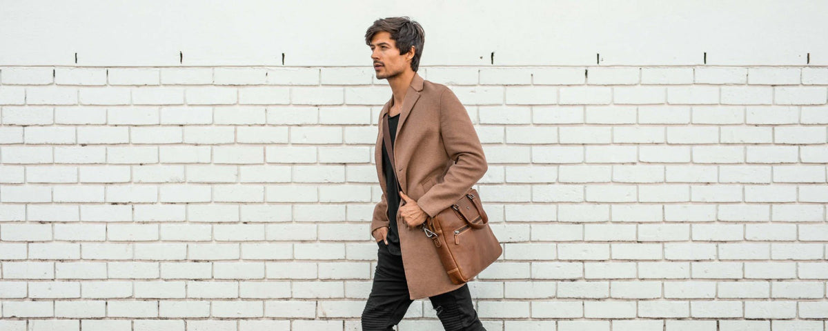 Man in a camel coat walking past a white brick wall with a brown leather messenger bag
