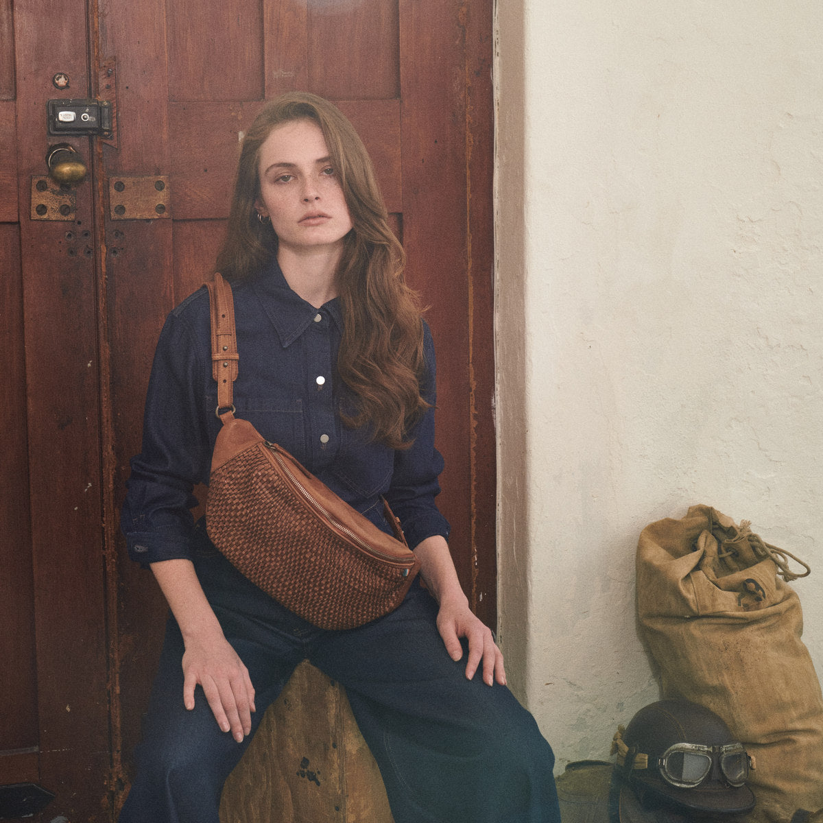 Cognac Bell Waist Bag worn crossbody by a woman seated on a crate in front of a wooden door