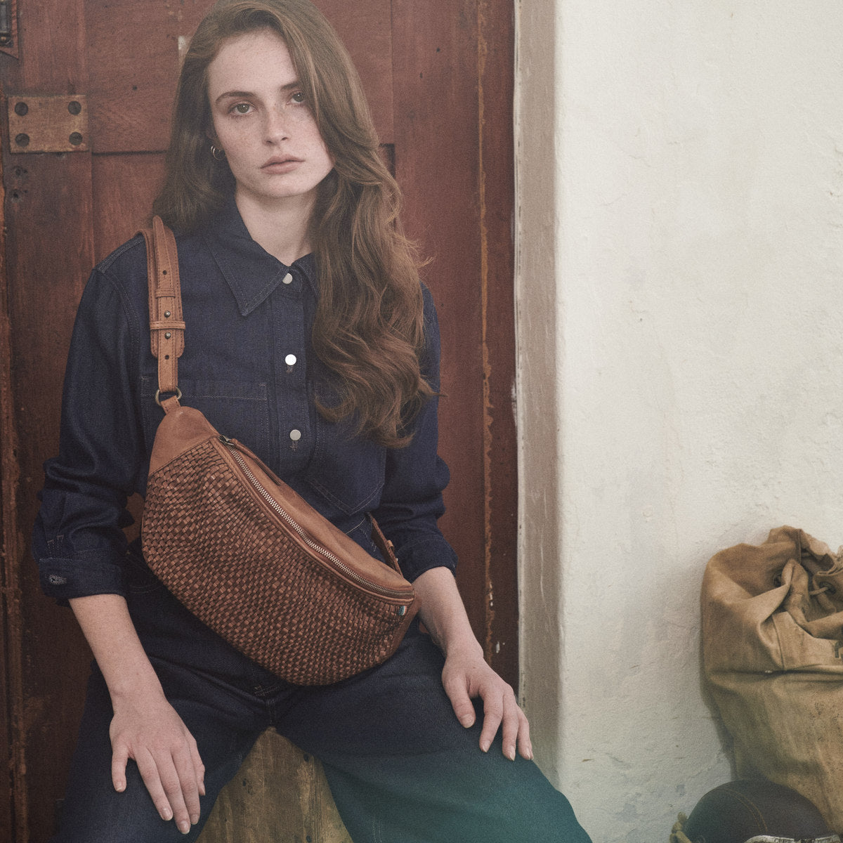 Bell cognac woven waist bag across a navy denim shirt, model seated by a wooden door
