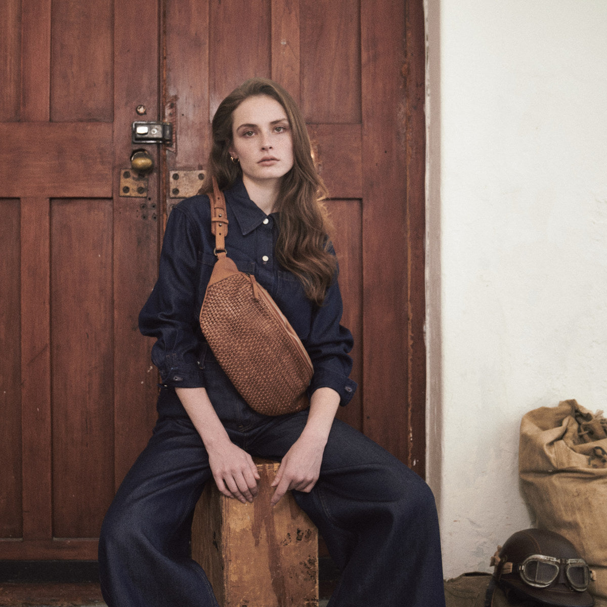 Woman seated by a wooden door wearing the cognac Bell Waist Bag, sandbags and goggles beside her