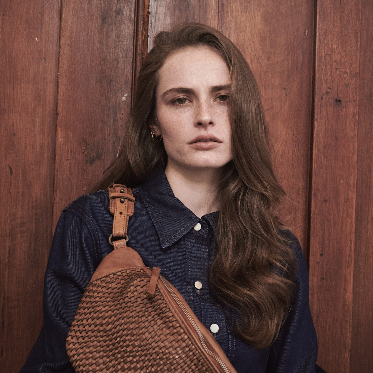 Bell Waist Bag in cognac worn over the shoulder, woman with long hair against a wooden door