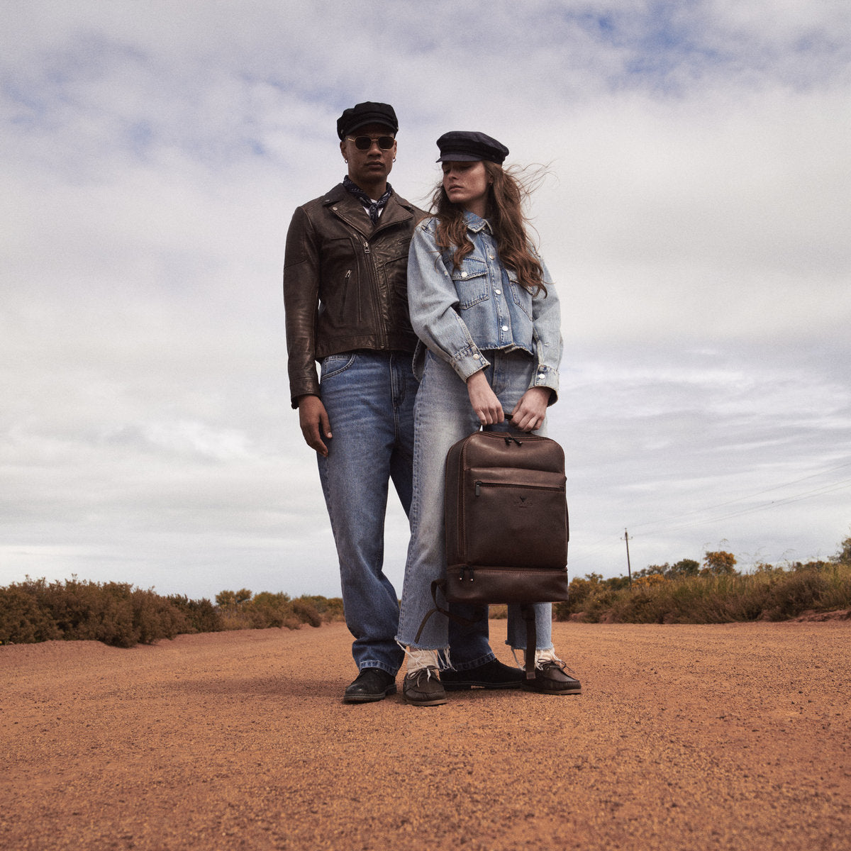 Couple on a dirt road, woman holding the brown James Laptop Backpack by its top handle