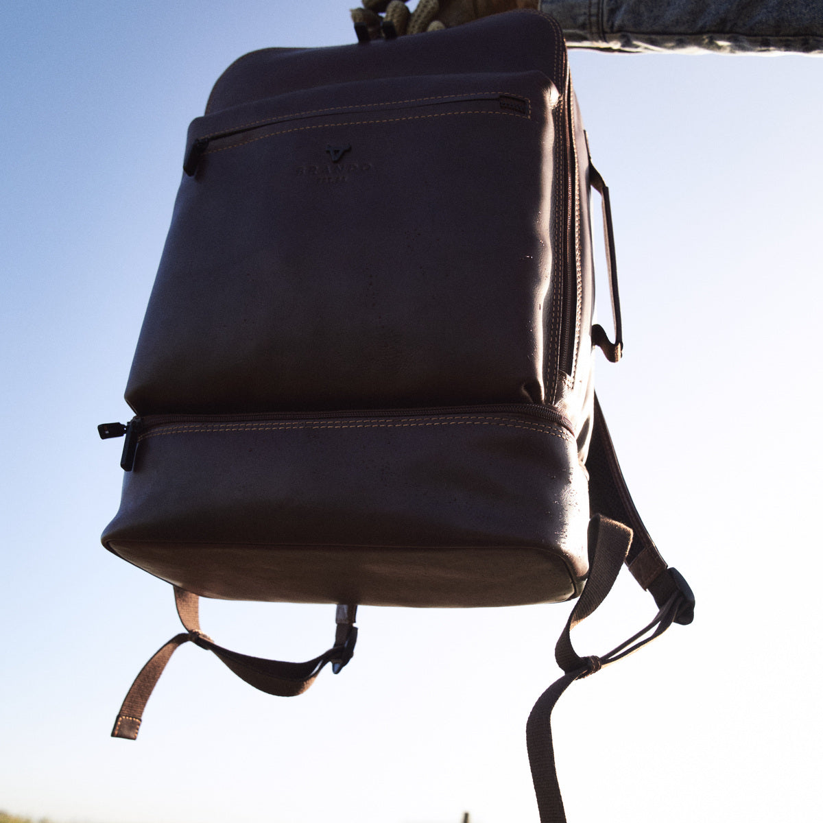 James Laptop Backpack in brown held up against the sky, front pocket and base compartment