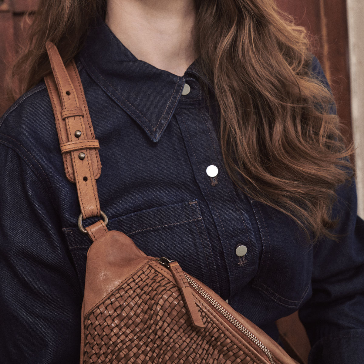 Close-up of the cognac Bell Waist Bag strap buckle and woven panel over a denim shirt