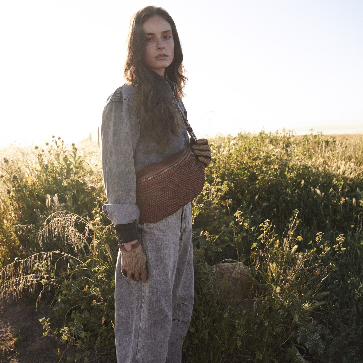 Bell Waist Bag in cognac worn crossbody by a woman in washed denim standing in long grass