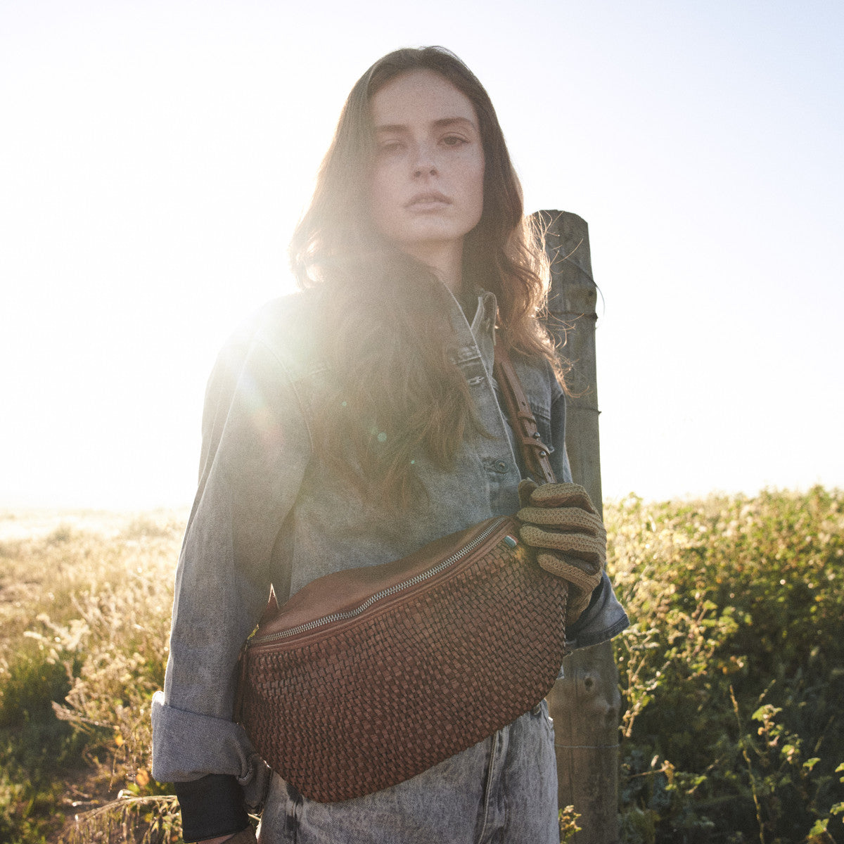 Cognac Bell Waist Bag across a washed denim jacket, woman by a fence post in morning sun