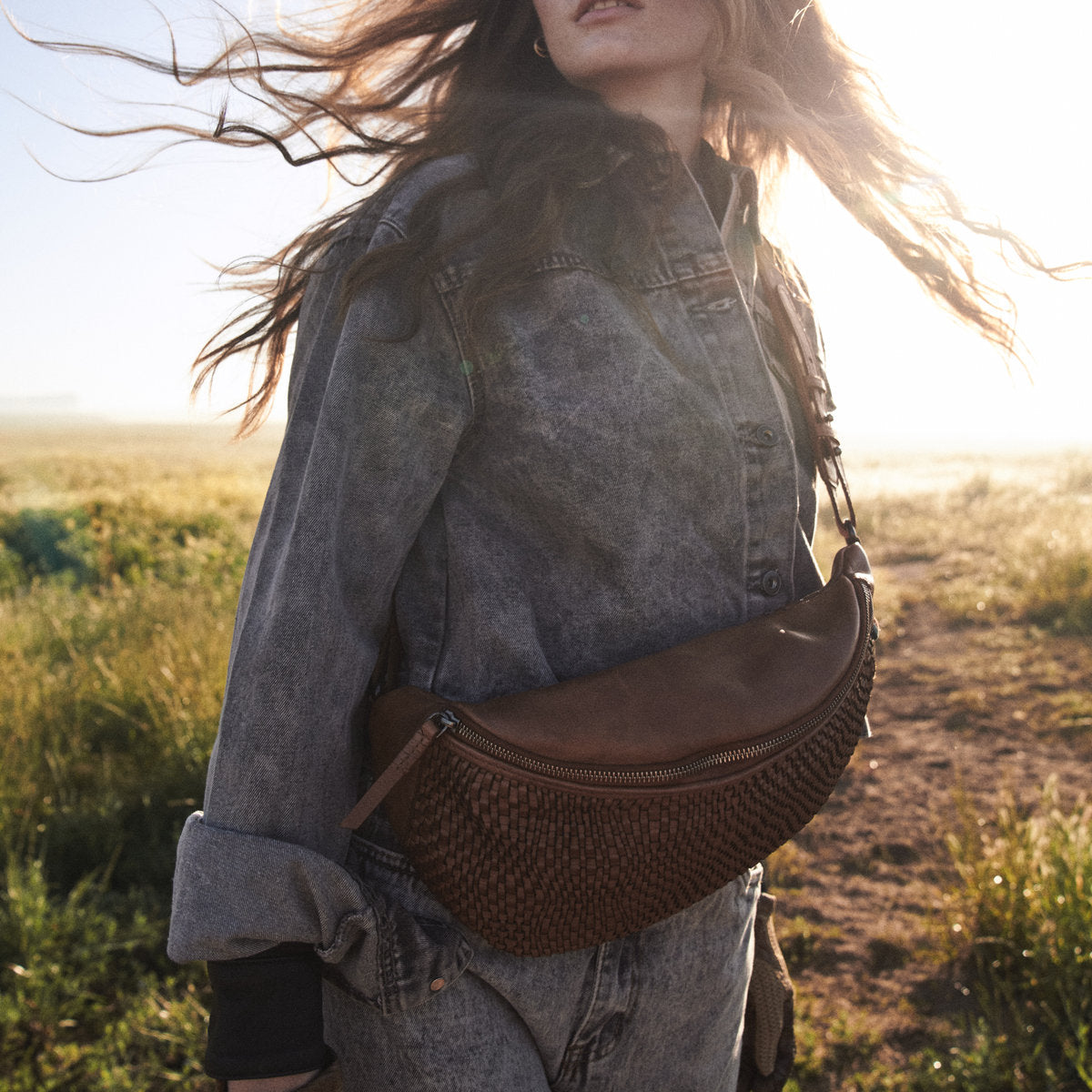 Bell Waist Bag in cognac worn across the body, wind in the model's hair on a farm track