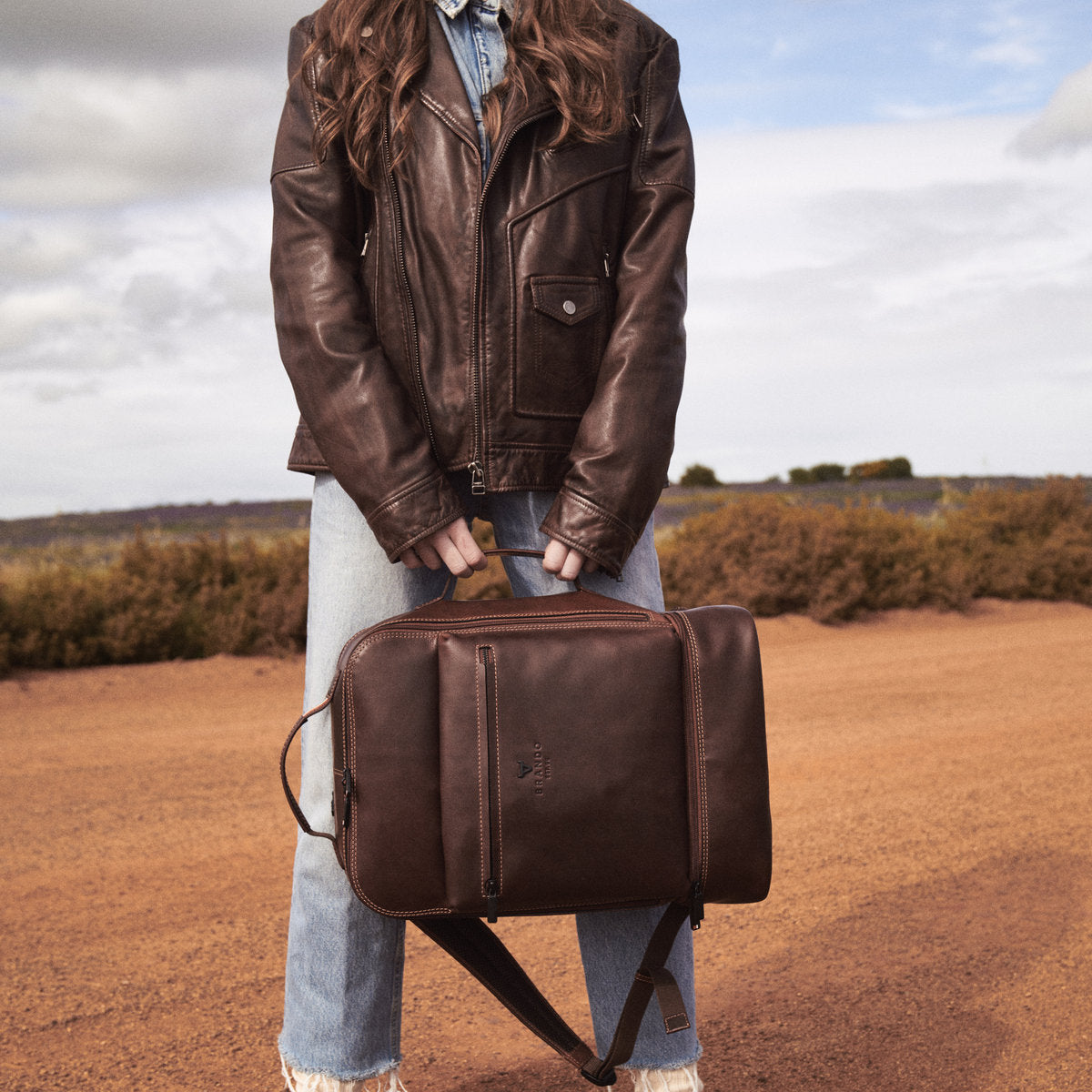Brown James Laptop Backpack held sideways by its side handle by a woman in a leather jacket