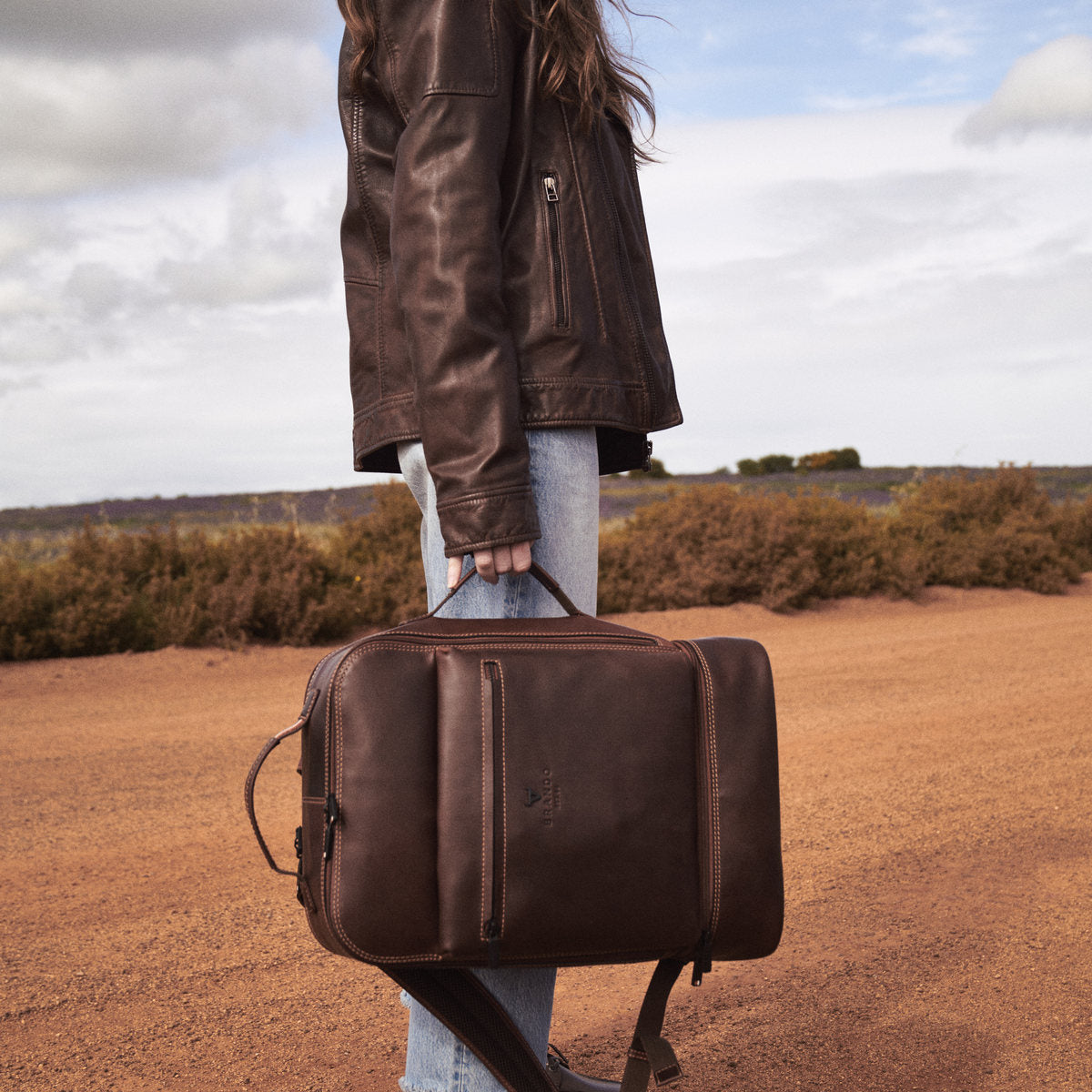 Side view of a woman holding the brown James Laptop Backpack by its side handle on a dirt road