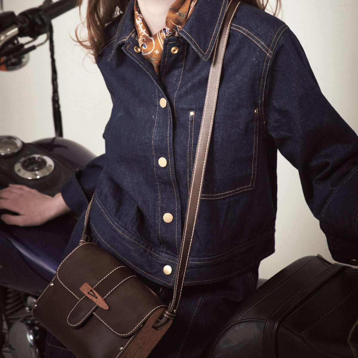 Brown Sally satchel with wooden toggle worn crossbody by a woman leaning on a motorbike