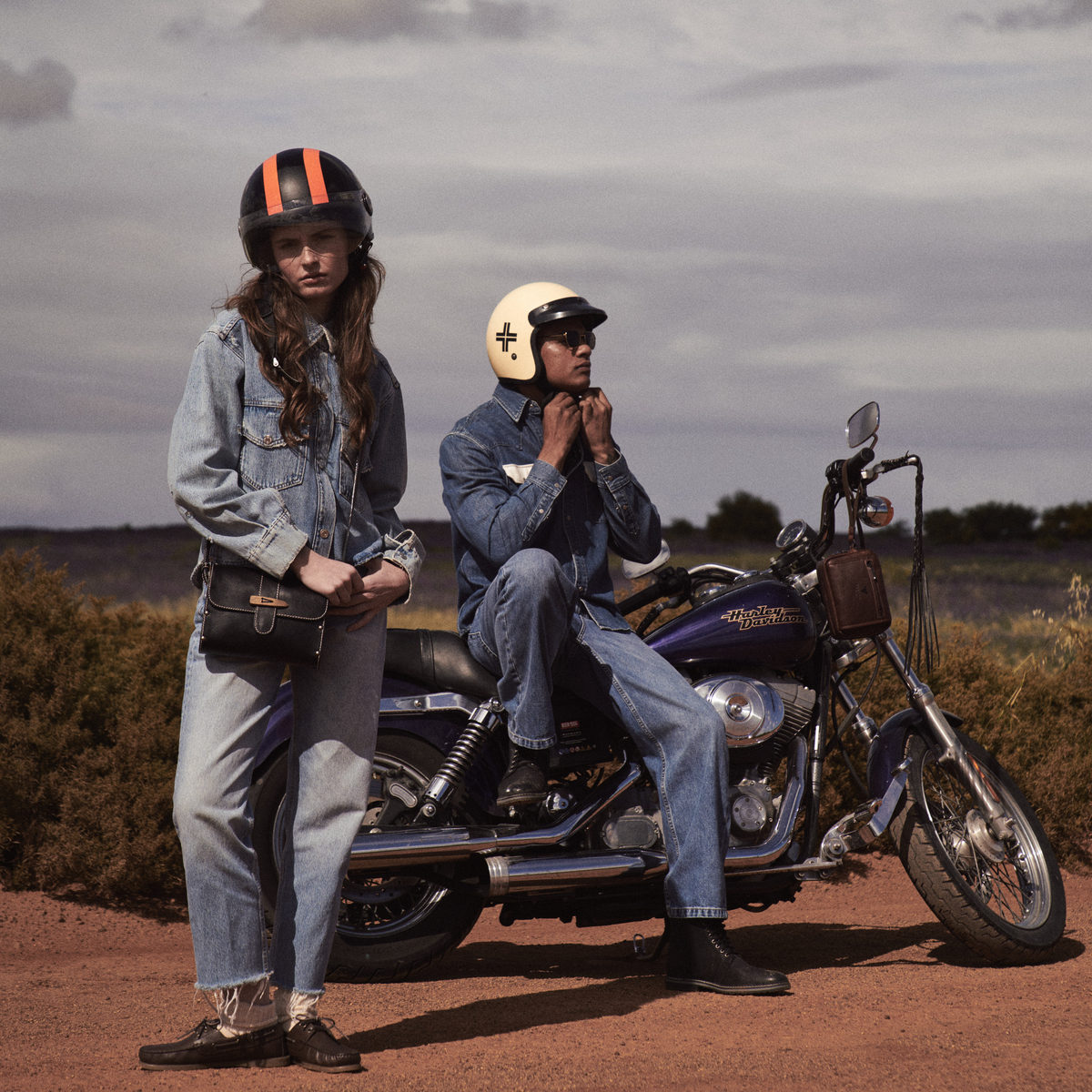 Woman wearing the black Sally satchel standing beside a rider on a purple motorbike on a dirt road