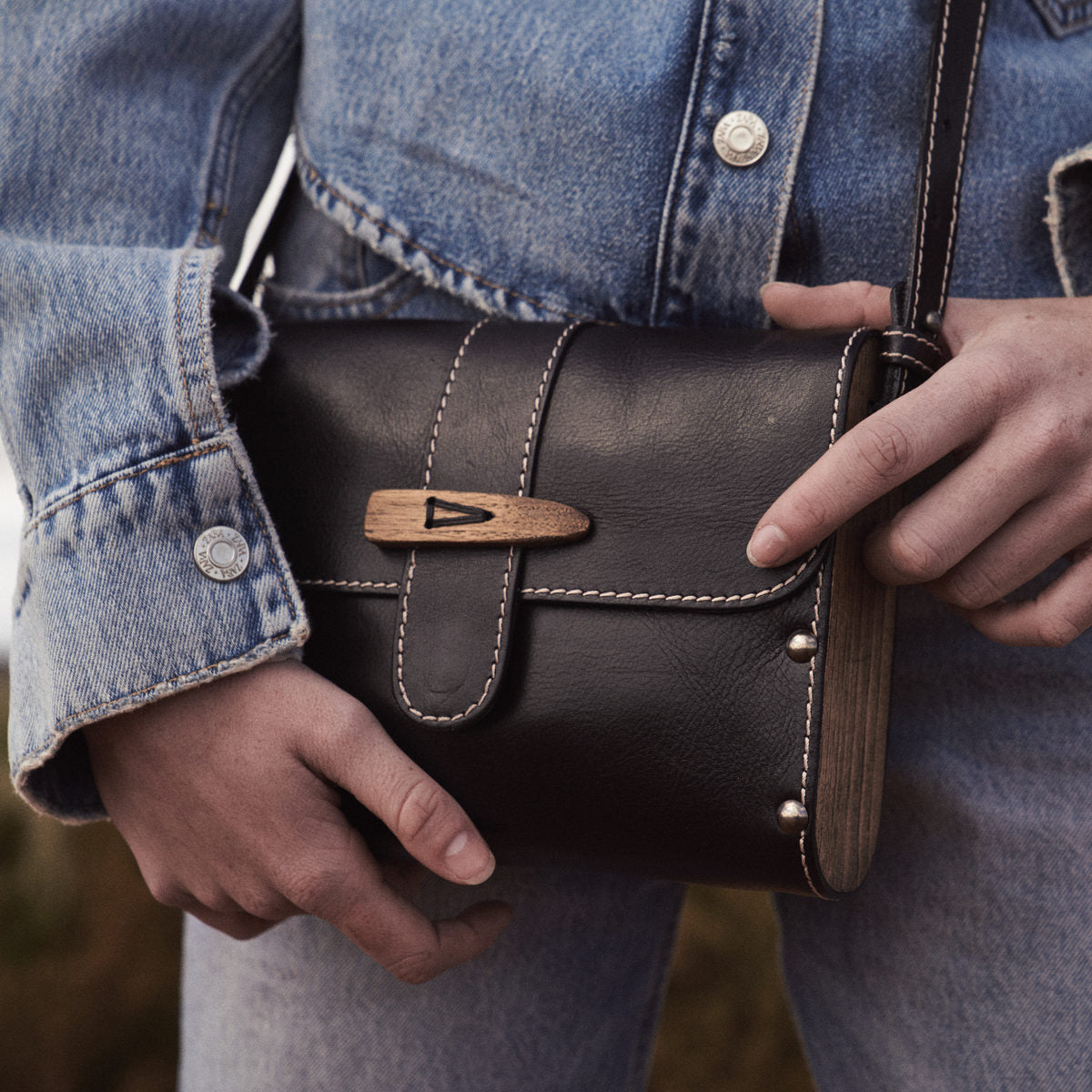 Close-up of the Sally satchel in black, wooden toggle and wood side panel held against denim